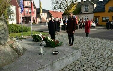 Dw&oacute;ch mężczyzn ubranych na czarno stoi przy pomniku, na kt&oacute;rym położone są kwiaty. Za nimi stoi kobieta ubrana na czerwono oraz grupa mężczyzn trzymająca sztandary. 