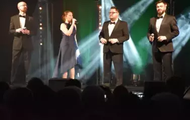 widzimy trzech pan&oacute;w i panią śpiewających na scenie 
