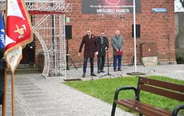 widzimy delegacje sztandarowe oraz trzech pan&oacute;w stojących za pomnikiem