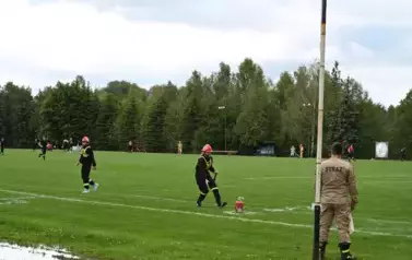 Dziewczyna z młodej drużyny strażackiej wykonuje zadanie konkurencyjne przy gaśnicy, za nią stoi chłopak r&oacute;wnież z drużyny strażackiej