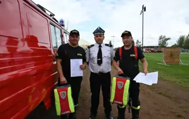 Strażacy stoją obok wozy strażackiego trzymając zaświadczenie o ukończeniu kursu oraz plecak z wyposażeniem medycznym