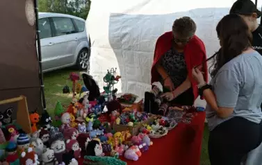 widzimy panią pokazującą przedmioty na stoisku oraz dwie panie patrzące na przedmioty