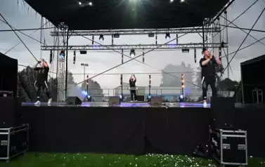 widzimy dw&oacute;ch pan&oacute;w śpiewających na scenie oraz pana stojącego w tle