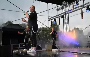widzimy dw&oacute;ch pan&oacute;w śpiewających na scenie oraz pana stojącego obok z telefonem w ręce 