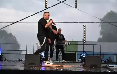 widzimy dw&oacute;ch pan&oacute;w śpiewających na scenie oraz pana stojącego w tle