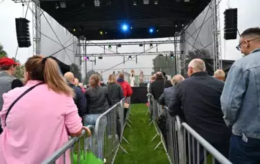 widzimy dużą grupę os&oacute;b słuchającą koncertu oraz osoby na scenie