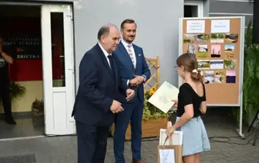 widzimy dw&oacute;ch pan&oacute;w przekazujących upominek dziewczynce