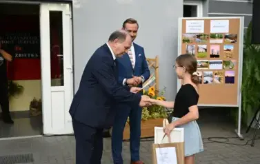 widzimy dw&oacute;ch pan&oacute;w przekazujących upominek dziewczynce