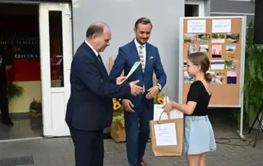 widzimy dw&oacute;ch pan&oacute;w przekazujących upominek dziewczynce