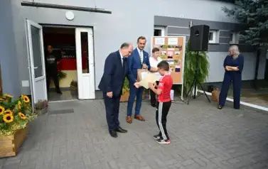 widzimy dw&oacute;ch pan&oacute;w przekazujących upominek chłopcu