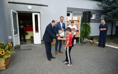 widzimy dw&oacute;ch pan&oacute;w przekazujących upominek chłopcu