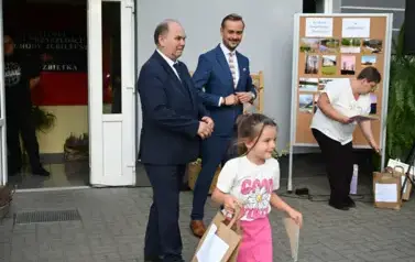 widzimy dziewczynke idącą z upominkami a w tle widzimy dw&oacute;ch pan&oacute;w
