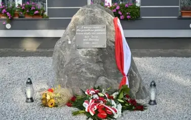 widzimy pomnik z zaczepioną polską flagą, kwiatami leżącymi na kamyszkach oraz dwoma zniczami obok 