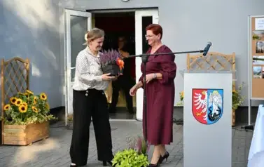 widzimy stojące dwie panie, jedna z nich trzyma kwiatka doniczkowego w ręce