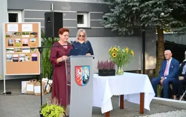 widzimy panią m&oacute;wiącą do mikrofonu oraz drugą panią stojącą w tle