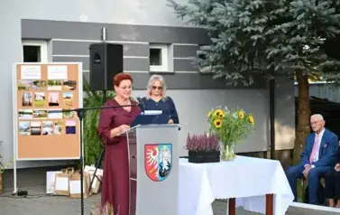 widzimy panią m&oacute;wiącą do mikrofonu oraz drugą panią stojącą w tle