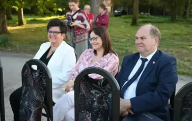 widzimy 2 panie oraz pana siedzących na krzesłach a w tle widzimy inne osoby
