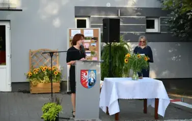 widzimy panią m&oacute;wiącą do mikrofonu oraz panią stojącą w tle