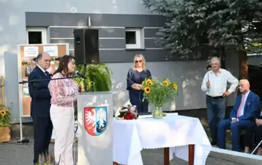 widzimy panią m&oacute;wiącą do mikrofonu oraz kilka osoby stojące oraz siedzące w tle 