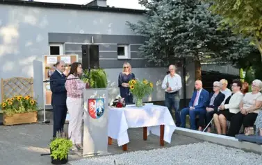 widzimy panią m&oacute;wiącą do mikrofonu oraz kilka osoby stojące oraz siedzące w tle 