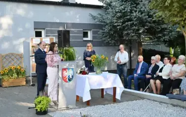 widzimy panią m&oacute;wiącą do mikrofonu oraz kilka osoby stojące oraz siedzące w tle 