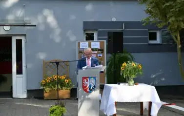 widzimy pana m&oacute;wiącego do mikrofonu oraz stojące obok słoneczniki w wazonie 