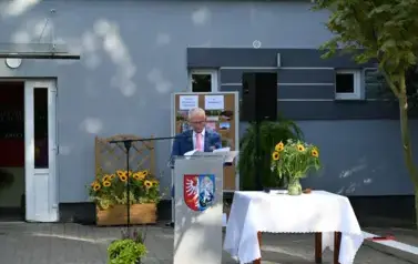 widzimy pana m&oacute;wiącego do mikrofonu oraz stojące obok słoneczniki w wazonie 