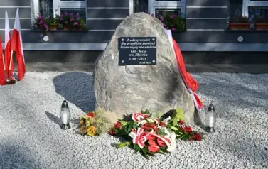 widzimy pomnik z zaczepioną polską flagą, kwiatami leżącymi na kamyszkach oraz dwoma zniczami obok 