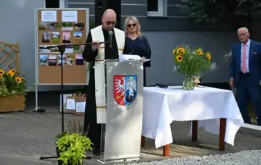 widzimy księdza m&oacute;wiącego do mikrofonu oraz panią stojącą za nim