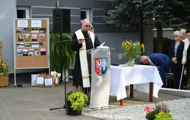 widzimy księdza m&oacute;wiącego do mikrofonu oraz panią stojącą za nim