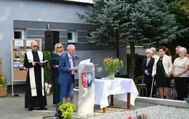 widzimy pana m&oacute;wiącego do mikrofonu oraz dwie osoby stojące za nim
