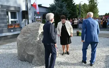widzimy 3 osoby stojące przed pomnikiem oraz liczną grupę os&oacute;b w tle