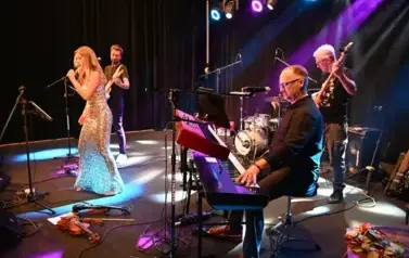 widzimy panią, kt&oacute;ra śpiewa do mikrofonu oraz pan&oacute;w grających na instrumentach