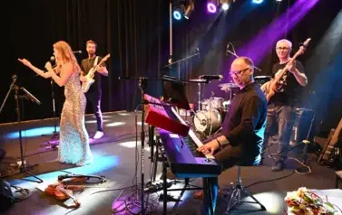 widzimy panią, kt&oacute;ra śpiewa do mikrofonu oraz pan&oacute;w grających na instrumentach