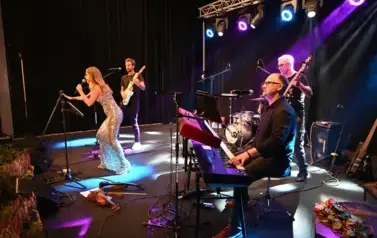 widzimy panią, kt&oacute;ra śpiewa do mikrofonu oraz pan&oacute;w grających na instrumentach