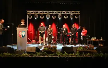 widzimy panią m&oacute;wiącą do mikrofonu, pana stojącego z teczką w ręce oraz grupe os&oacute;b stojącą na scenie