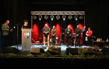 widzimy panią m&oacute;wiącą do mikrofonu, pana stojącego z teczką w ręce oraz grupe os&oacute;b stojącą na scenie