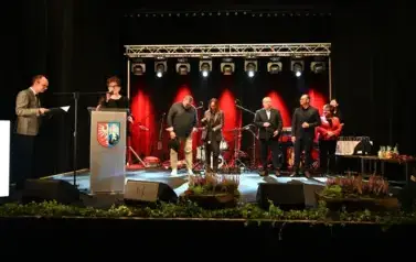 widzimy panią m&oacute;wiącą do mikrofonu, pana stojącego z teczką w ręce oraz grupe os&oacute;b stojącą na scenie