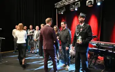 widzimy pana podającego podarunek grukie os&oacute;b stojących w rzędzie na scenie