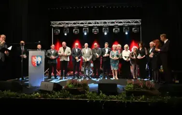 widzimy pana m&oacute;wiącego przez mikrofon oraz grupe os&oacute;b stojącą na scenie