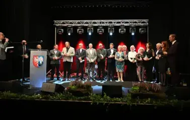 widzimy pana m&oacute;wiącego przez mikrofon oraz grupe os&oacute;b stojącą na scenie