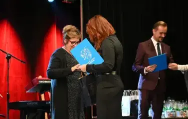 widzimy dwie pani, jedna z nich gratuluje drugiej pani oraz przekazuje jej nagrode
