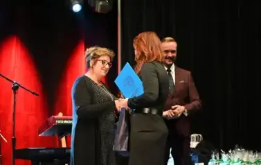 widzimy dwie pani, jedna z nich gratuluje drugiej pani oraz przekazuje jej nagrode