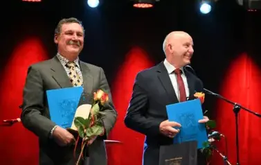 widzimy dw&oacute;ch pan&oacute;w uśmiechających się oraz trzymających w rękach r&oacute;że oraz teczke