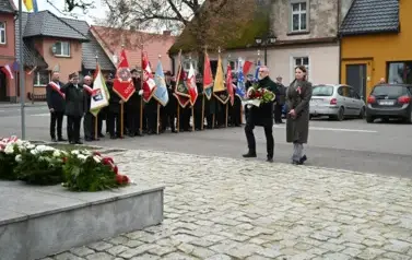 widzimy pana i panią idących z wieńcem w strone pomnika, w tle widoczne są sztandary