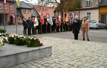 widzimy 2 panie idące z wieńcem w strone pomnika, w tle widoczne są sztandary