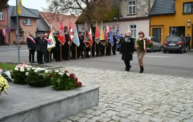 widzimy 2 panie idące z wieńcem w strone pomnika, w tle widoczne są sztandary