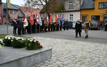 widzimy 2 panie idące z wieńcem w strone pomnika, w tle widoczne są sztandary