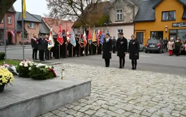 widzimy pana i 2 panie stojące przed pomnikiem, w tle widoczne są sztandary