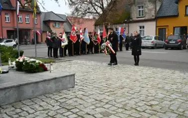 widzimy panią i pana idącego z wieńcem w strone pomnika, w tle widoczne są sztandary